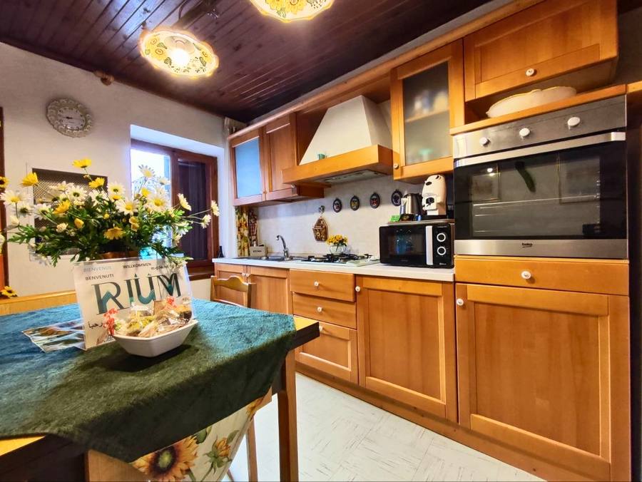Cucina in legno di Rium Dolomiti con tavolo, insegna benvenuto, forno, microonde e fiori gialli